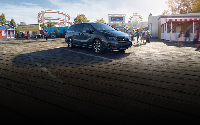 2026 Honda Odyssey Elite in Smoke Blue Pearl front parked on a boardwalk near a county fair (Minivan).