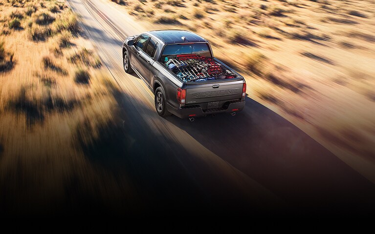 2026 Honda Ridgeline RTL in Modern Steel Metallic back driving on a desert road with a loaded bed (Midsize Pickup Truck).