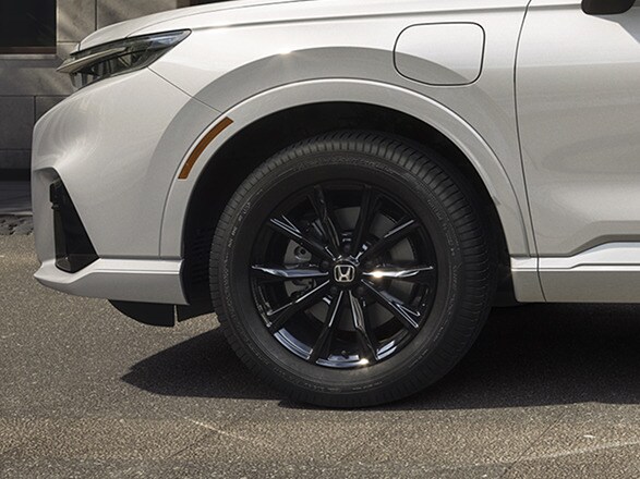 Wheel detail of 2026 CR-V e:FCEV in Platinum White Pearl showing 18" alloy wheels parked in a clean modern urban location, with trees / greenery.