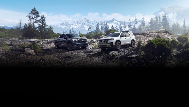2026 Pilot in Platinum White Pearl next to a 2026 Ridgeline with HPD package in Crystal Black Pearl, parked in an outdoor terrain surrounded by trees.